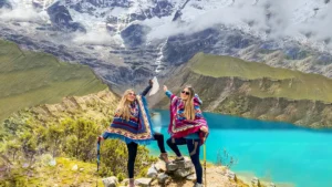 Two female travelers wearing colorful ponchos posing above the turquoise Humantay Lake during the Salkantay Pass Trek 2 Days.