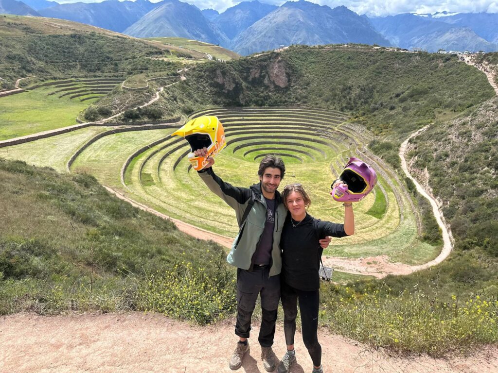 Couple holding biking helmets overlooking the circular terraces of Moray in the Sacred Valley, Peru.