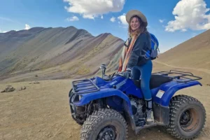 Smiling traveler riding a blue ATV near Rainbow Mountain (Vinicunca) in Cusco, Peru with colorful Andean hills in the background.