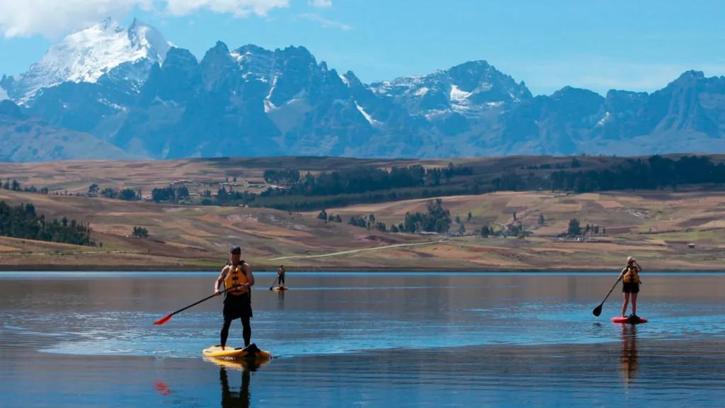Travelers practicing stand up paddle on Huaypo Lake with snow-capped Andes mountains in the background, Cusco Peru.